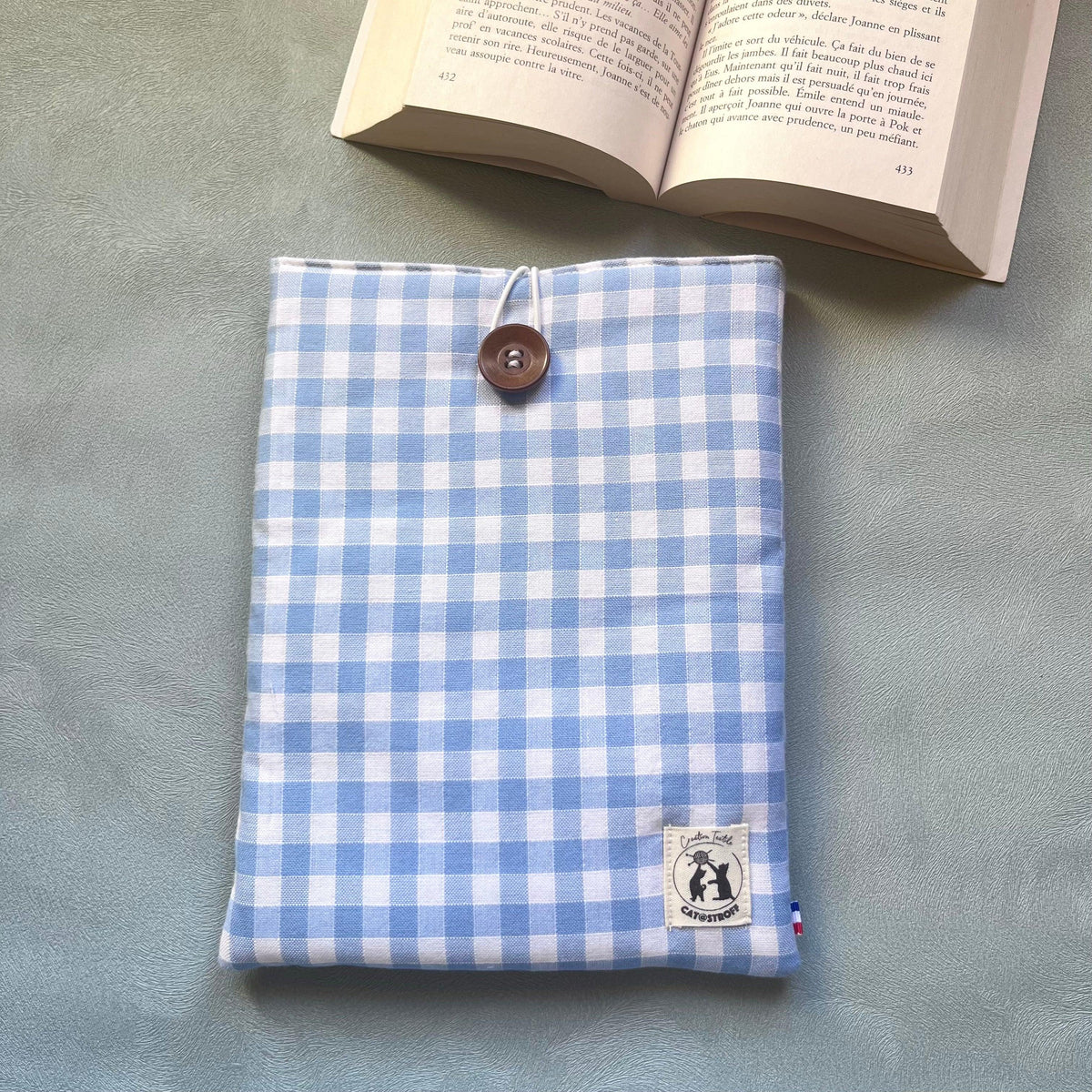 Pochette à livre vichy bleu posée à plat avec un roman ouvert, bouton bois et étiquette Catastroff visibles, vue de dessus