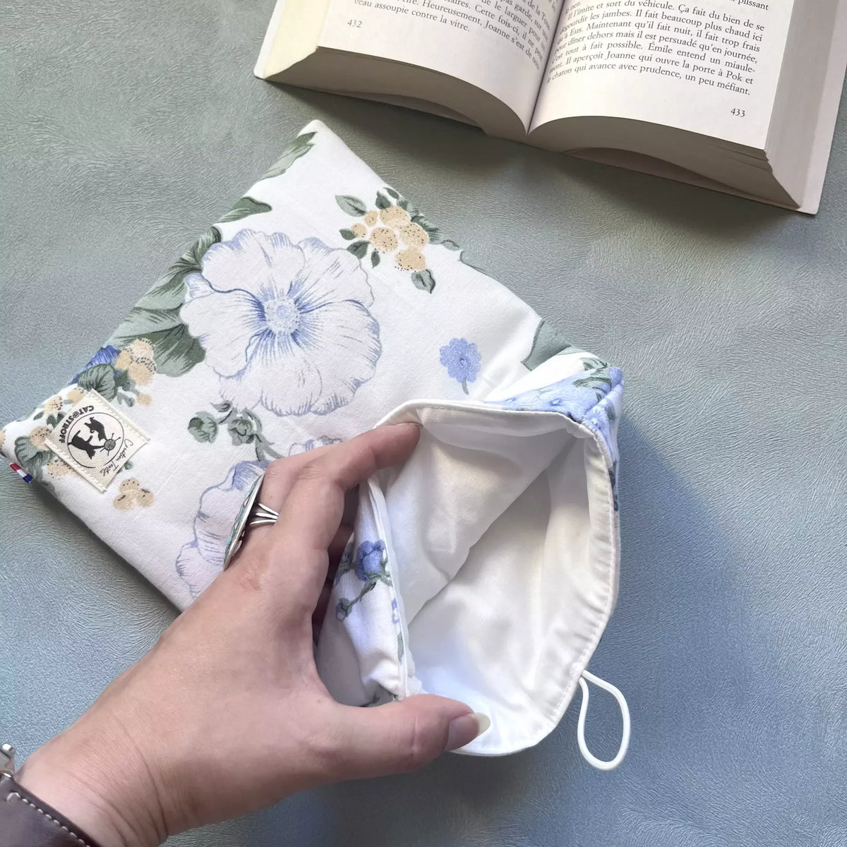 Main ouvrant la pochette à livre draps fleuri pour montrer l'intérieur blanc propre et le rembourrage molleton, livre ouvert en fond