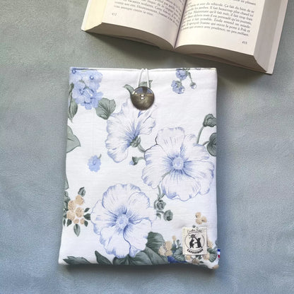 Pochette à livre draps fleuri posée à plat avec un livre ouvert en arrière-plan, motif floral bleu et blanc, bouton chiné visible, étiquette Catastroff