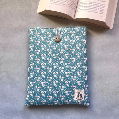Pochette à livre bleu paquerette Catastroff posée à plat, motif pâquerettes blanches sur fond bleu canard, bouton chiné et livre ouvert en arrière-plan.