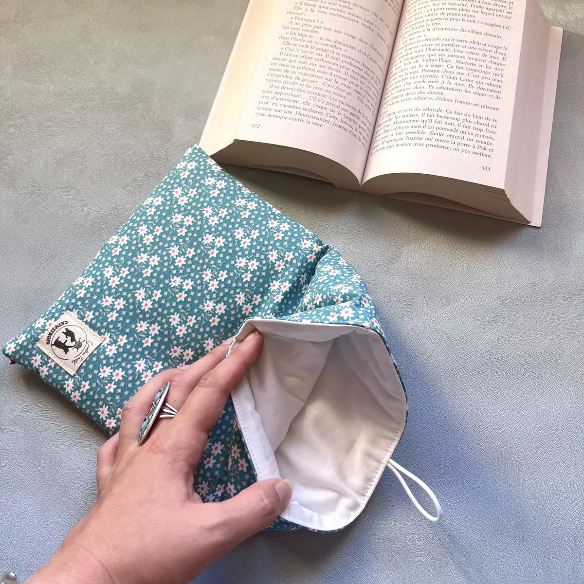 Main ouvrant la pochette protège-livre tissu fleurs Catastroff, dévoilant la doublure intérieure blanc crème et le rembourrage molleton épais.