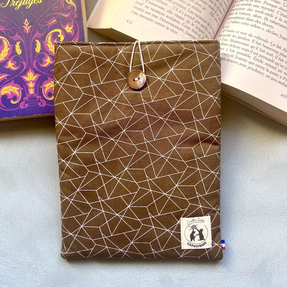 Pochette à livre en coton marron motif géométrique blanc, fermée par un bouton nacré de récupération, posée ici sur un livre ouvert cousue main par Catastroff