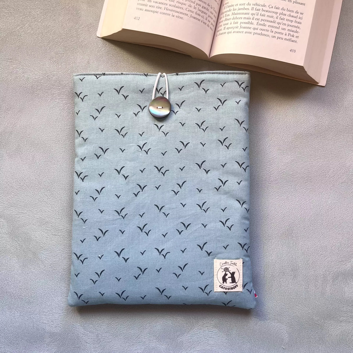 Pochette à livre bleu oiseau Envolée Bleue vue de face posée à plat, motif oiseaux en vol sur coton bleu ciel, étiquette Catastroff visible