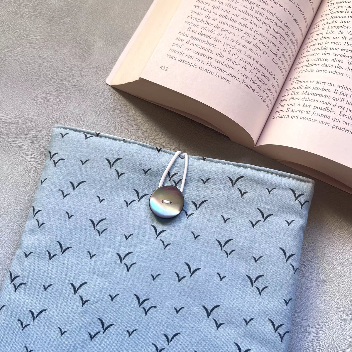 Gros plan sur la fermeture élastique blanc et bouton nacré de récupération de la pochette à livre bleu oiseau, livre ouvert en arrière-plan