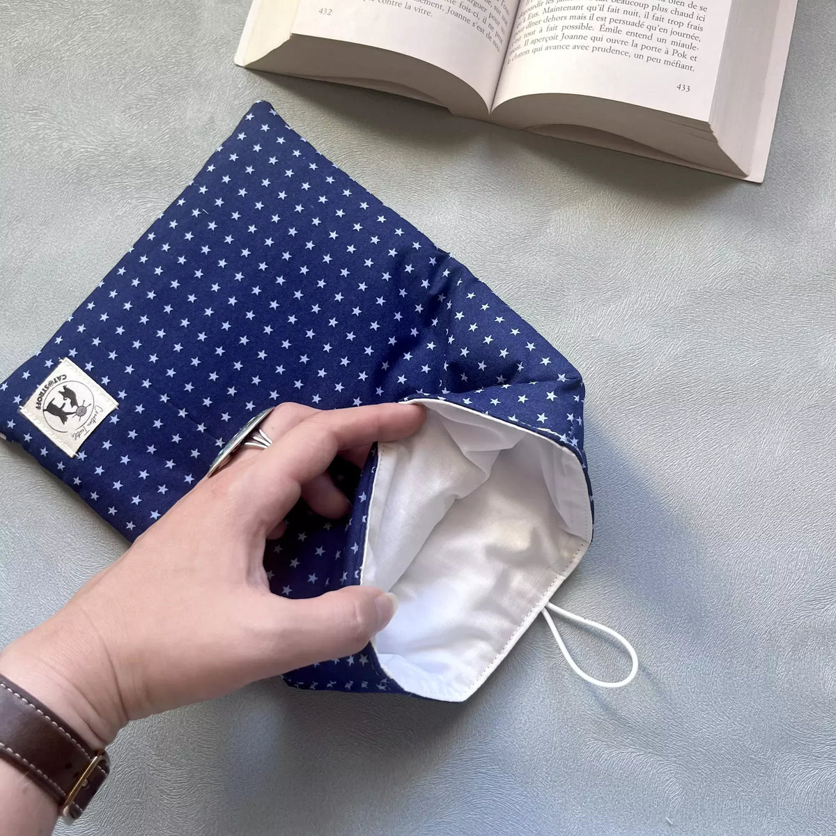 Main ouvrant la pochette protège livre de poche fait main La Belle Étoile, révélant la doublure intérieure coton clair et l'élastique blanc de fermeture.