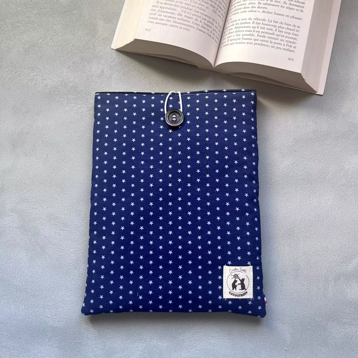 Pochette à livre bleu etoilé La Belle Étoile posée à plat, motif étoiles claires sur fond bleu nuit, bouton de récupération visible, livre ouvert en arrière-plan.