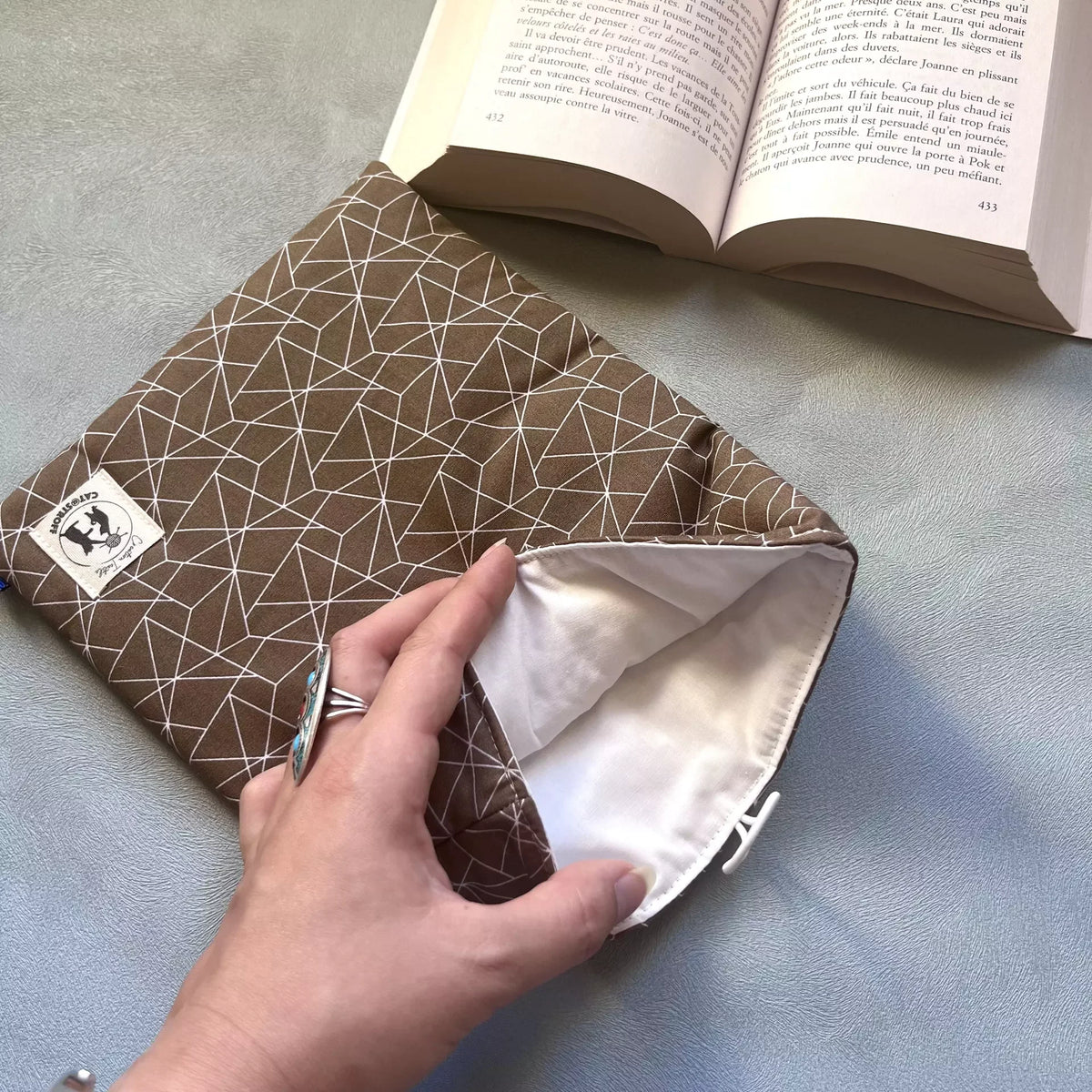 Main ouvrant la pochette à livre Moka Graphique Catastroff, révélant la doublure intérieure en coton naturel clair, tissu extérieur coton marron géométrique