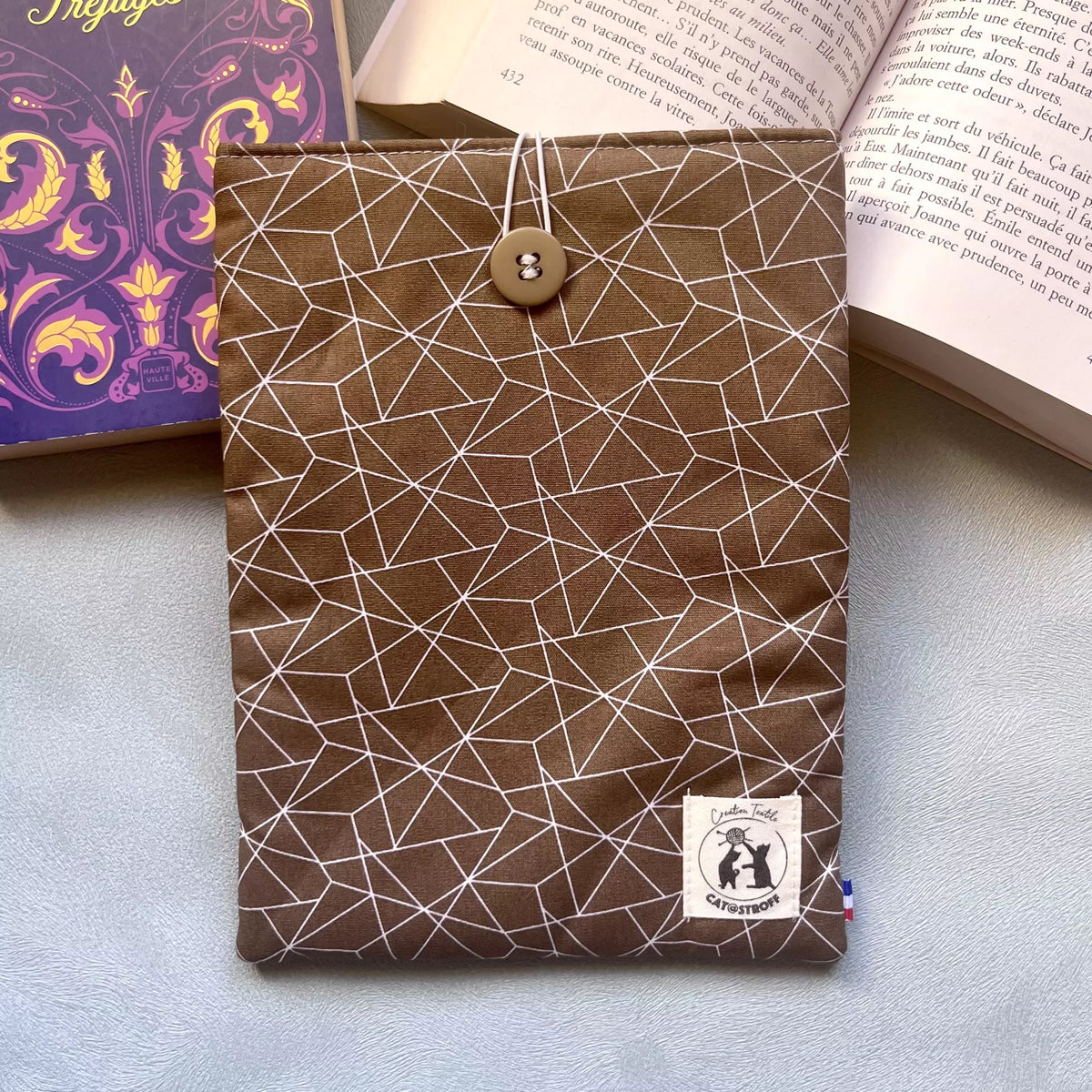 Housse livre poche voyage tissu Puzzle Café posée à plat entre deux livres ouverts, motif géométrique brun et blanc, étiquette Catastroff visible.
