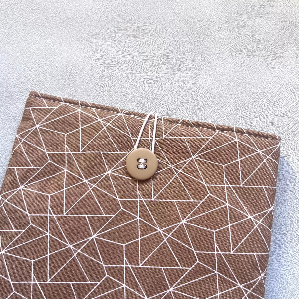 Gros plan sur la fermeture élastique et le bouton de récupération de la pochette livre de poche en tissu coton marron ligné Catastroff.