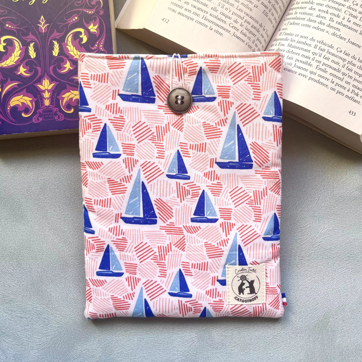 Pochette à livre tissu coton imprimé voiliers bleus et traits roses, cousue main, fermée par un bouton récupéré et posé sur un livre ouvert 