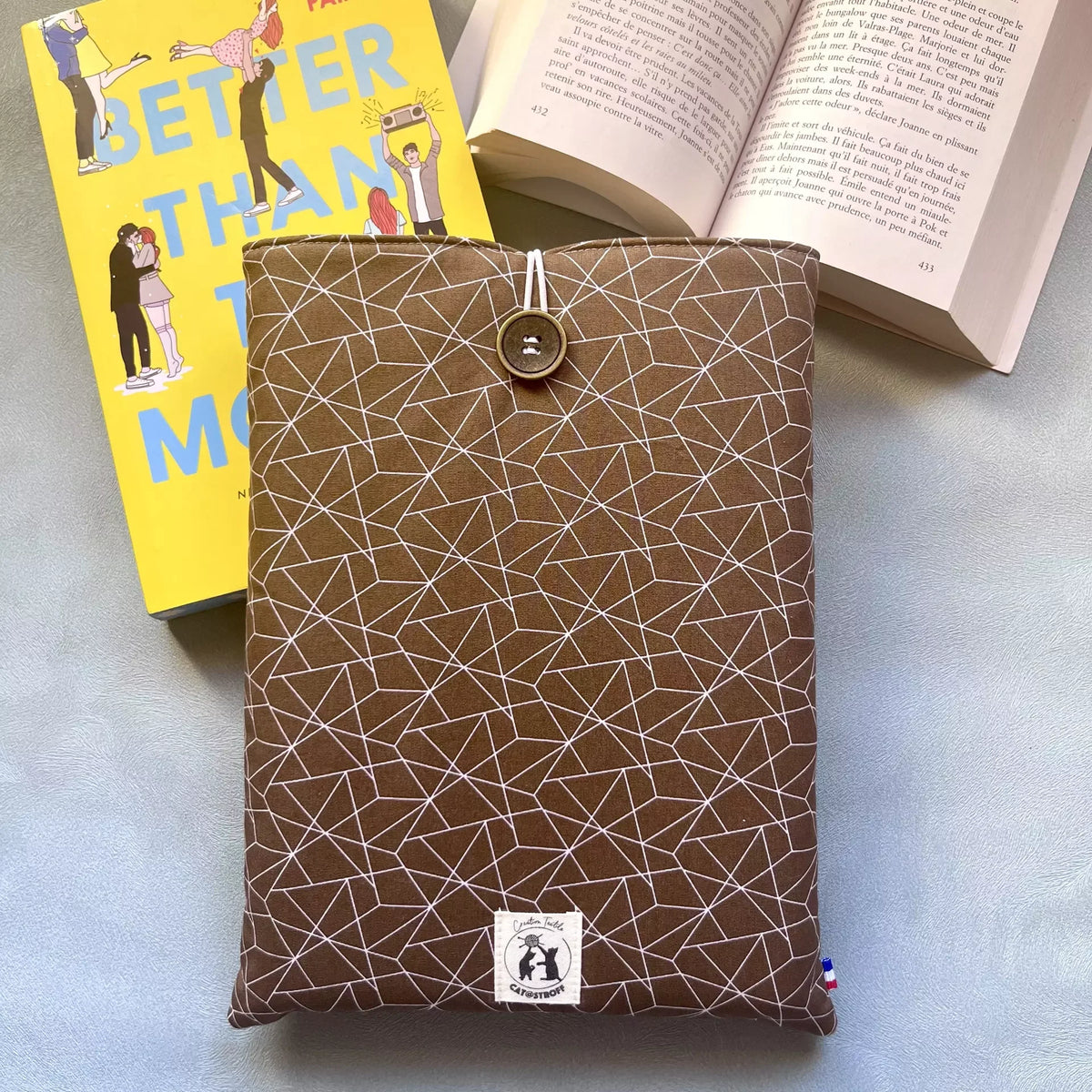 Pochette à livre grand format Cacao Graphique posée avec un roman broché et un livre ouvert, illustrant son usage au quotidien