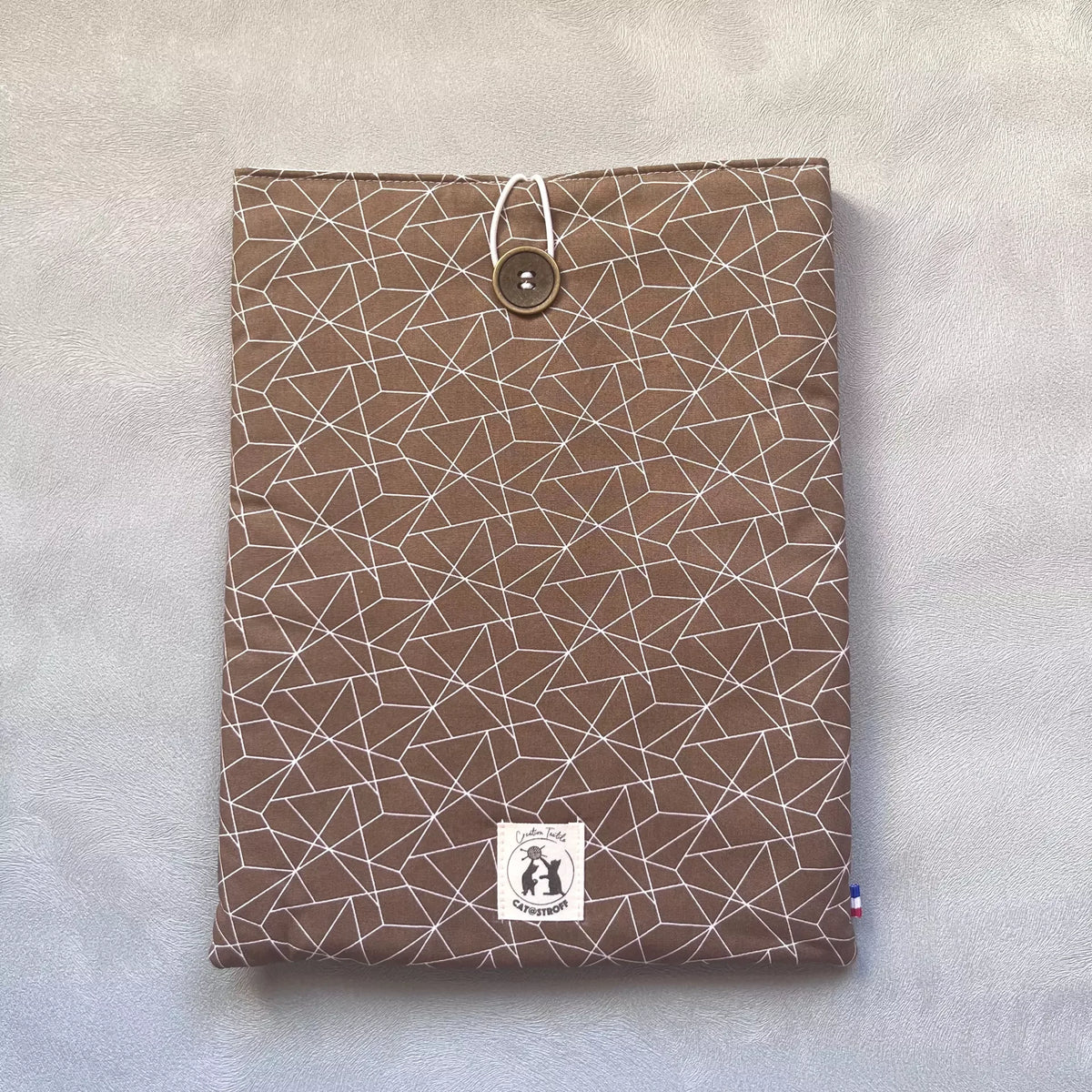 Pochette livre broché marron vue de face fermée, motif triangles géométriques blancs, bouton de récupération ton bois et étiquette Catastroff