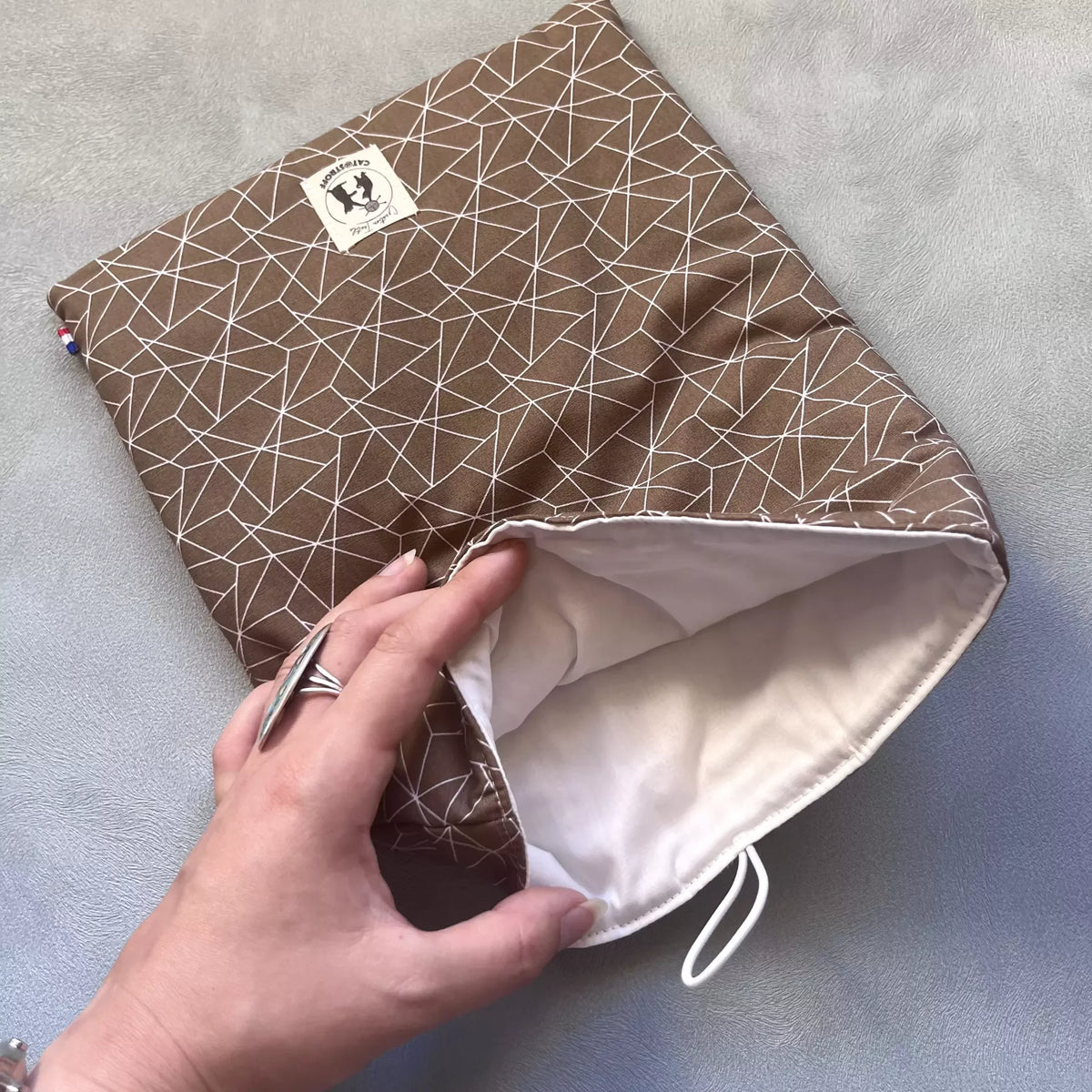 Pochette à livre grand format Cacao Graphique ouverte, doublure claire visible, tenue en main pour montrer la profondeur et le molleton intérieur
