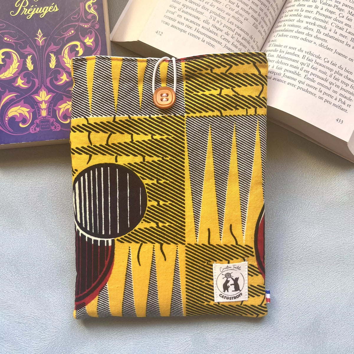 Pochette livre de poche en tissu wax africain jaune et noir avec bouton caramel, ici posée sur un livre ouvert, faite main par Catastroff